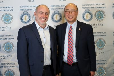 Two men stand in front of a backdrop with sponsor logos at the 2024 National Prosecution Best Practices Conference.