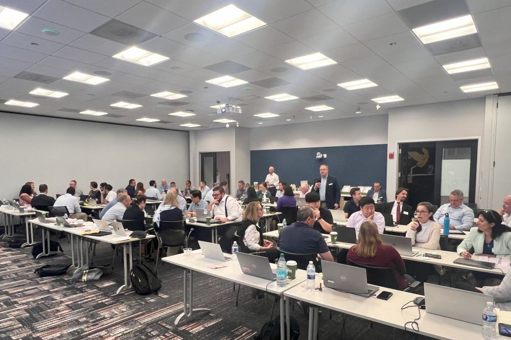 Multiple rows of people sit at long tables in a discussion at the Microsoft PCE Hackathon. 