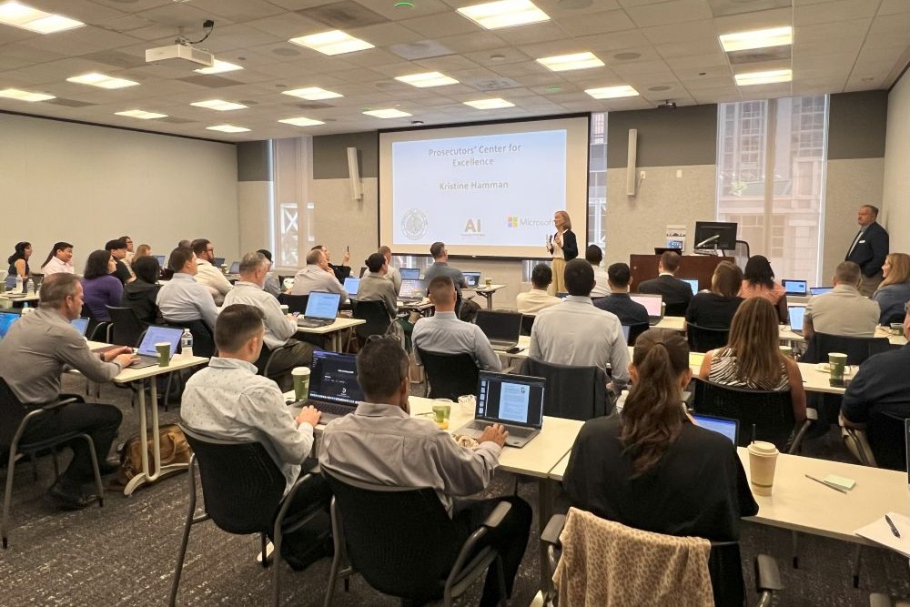 Prosecutors' Center for Excellence Executive Director Kris Hamman stands at the front of a classroom at the Microsoft PCE Hackathon with a presentation on the projector screen behind her. Rows of people face her while sitting at long tables. 