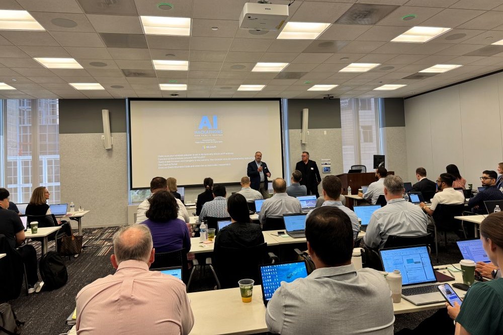 Two speakers stand at the front of a large room with rows of people on laptops in the audience. 