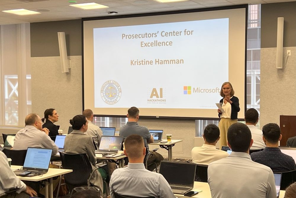 Prosecutors' Center for Excellence Executive Director Kris Hamman stands at the front of a classroom at the Microsoft PCE Hackathon with a presentation on the projector screen behind her. Rows of people face her while sitting at long tables. 