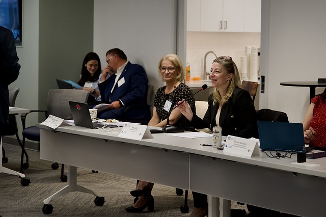 Two people sit at a long white table at the PERF Prosecutor Police Collaboration Meeting. Two people sit behind them in chairs against the wall. 