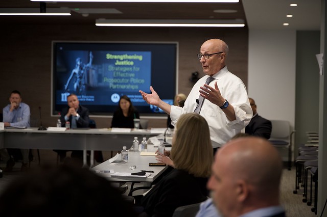 A speaker stands with hands raised at the PERF Prosecutor Police Collaboration Meeting. Others seated at nearby tables sit listening to the speaker. 