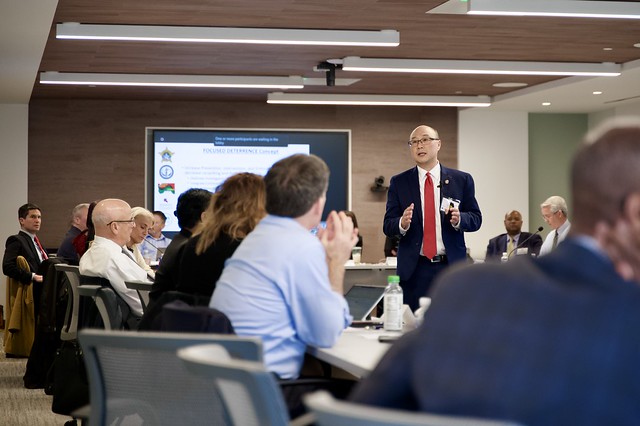 A speaker wearing a black jacket and red tie stands with his hands out as a line of people seated at a desk look at him. 