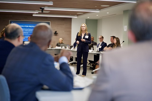 A speaker in a black suit stnds in the middle of a classroom surrounded by long tables filled with seated people at the PERF Prosecutor Police Collaboration Meeting.